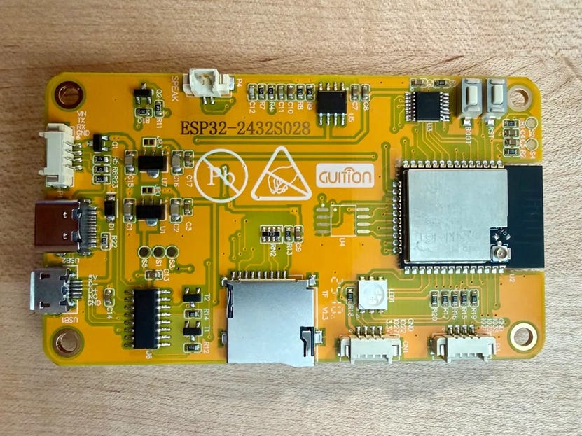 Close-up photo of a yellow ESP32-2432S028 microcontroller board placed on a wooden surface, showing ports, chips, and connectors.