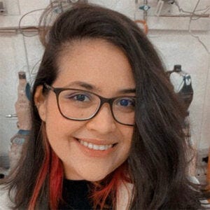 Mulher, cabelos castanhos com mechas rosas. Usa óculos e jaleco.