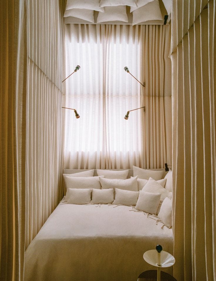 This may contain: a white bed sitting under a window next to a table with a lamp on top of it