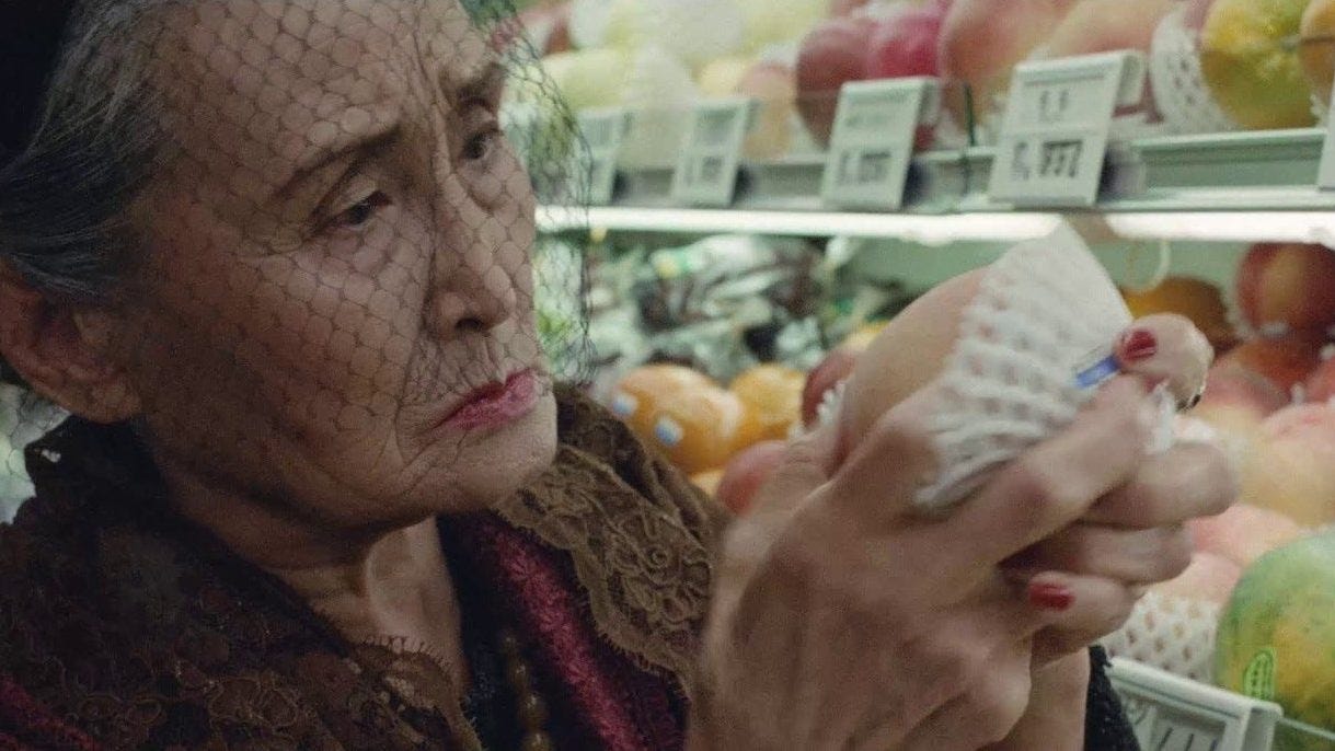 An elderly woman with a thoughtful expression examines fruit wrapped in foam netting at a supermarket. She wears a brown coat and a black veil.