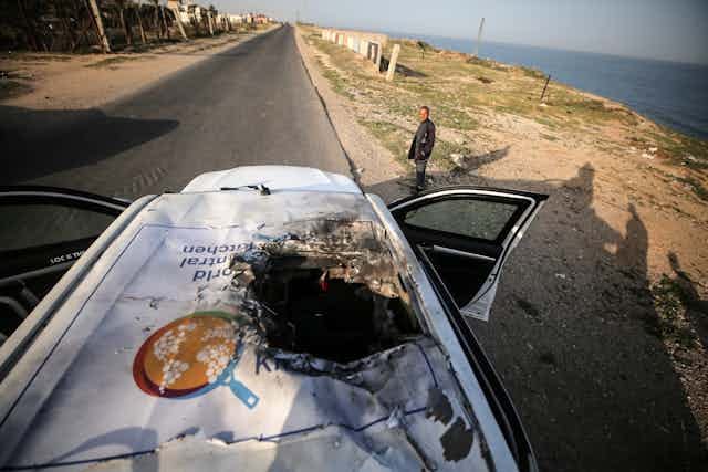 A man stands near a white vehicle that has a large hole in its top and its doors open. The road is empty and the ocean is nearby. A man stands near a white vehicle that has a large hole in its top and its doors open. The road is empty and the ocean is nearby.