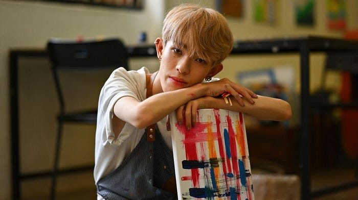 Nonbinary teen in a white t-shirt with arms crossed, propped on an abstract painting.