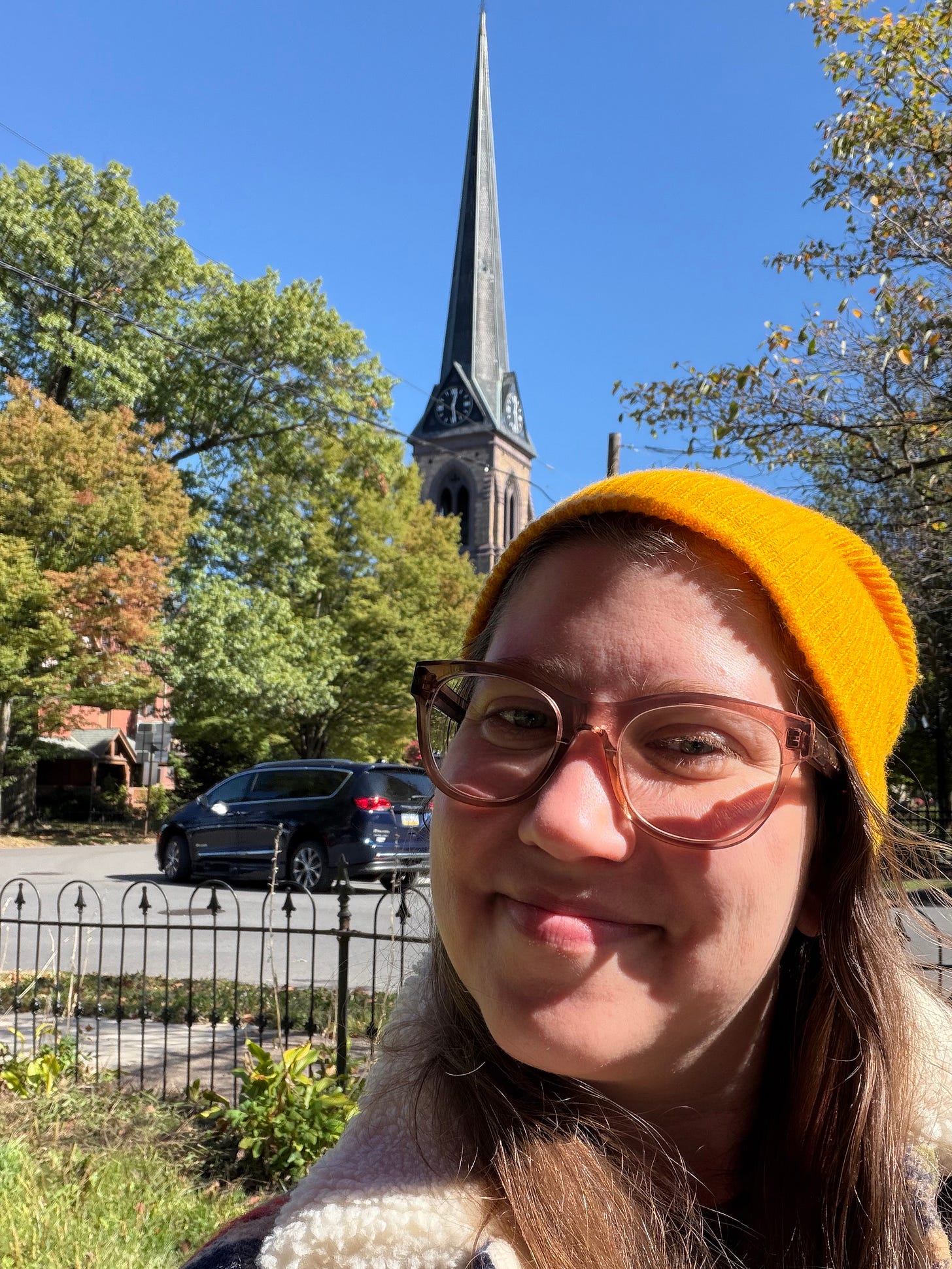A woman wears glasses and a beanie in front of leaves, a car, and a large church in the back across the street