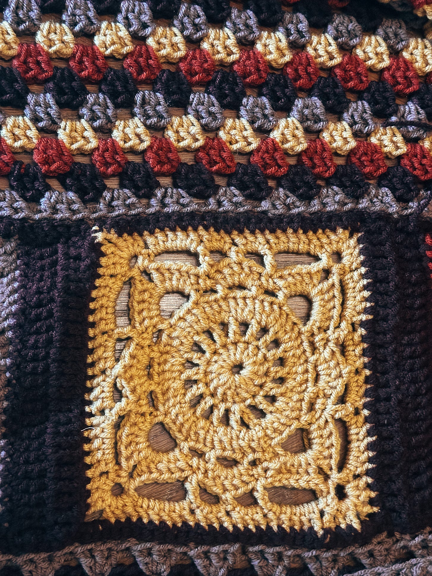 close-up of a granny square stitch, rows of alternating brown, mauve, yellow, and burnt orange, in the bottom half of the image is a large willow square in yellow, a mandala type pattern close-up of a granny square stitch, rows of alternating brown, mauve, yellow, and burnt orange, in the bottom half of the image is a large willow square in yellow, a mandala type pattern