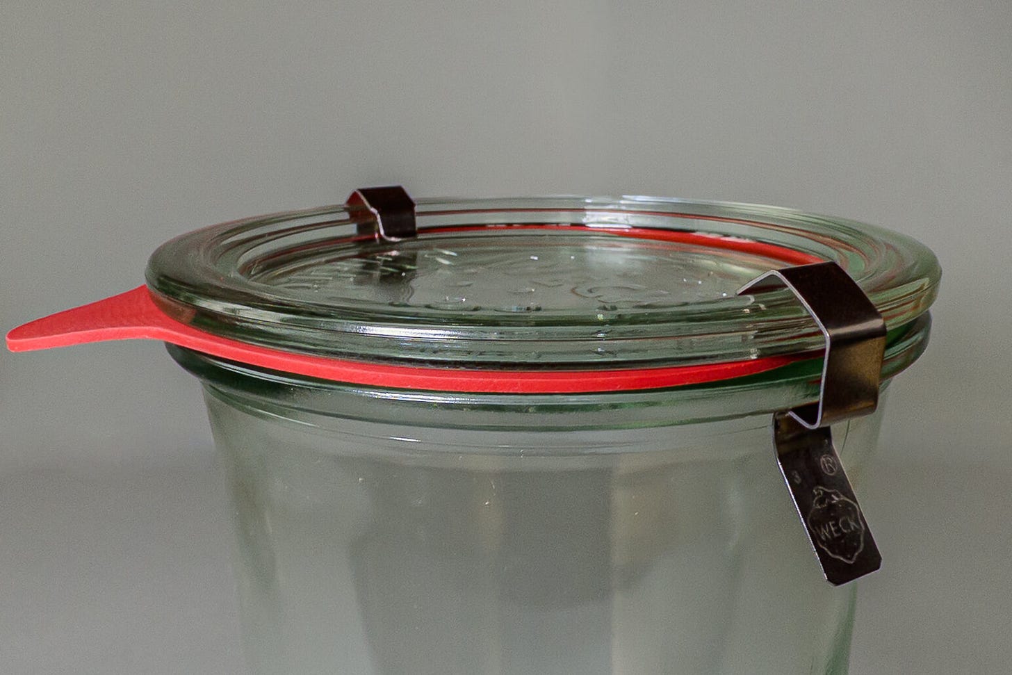 Detailaufnahme eines WECK-Rundrandglases mit Glasdeckel und Metallklammern; der orange Gummiring liegt zwischen Deckel und Glas und dichtet den charakteristischen Rundrand luftdicht ab. Detailaufnahme eines WECK-Rundrandglases mit Glasdeckel und Metallklammern; der orange Gummiring liegt zwischen Deckel und Glas und dichtet den charakteristischen Rundrand luftdicht ab.