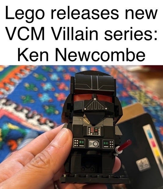 Text above a photo: "Lego releases new VCM Villain series: Ken Newcombe". Below, a hand holds a Lego BrickHeadz style Darth Vader figure with a red lightsaber over a colorful rug.