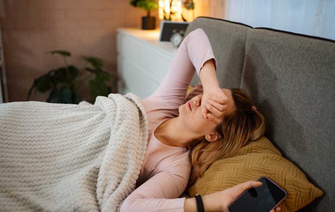 Woman laying in bed in soft morning light with one arm over her eyes, holding her phone in the other hand, struggling to wake up.