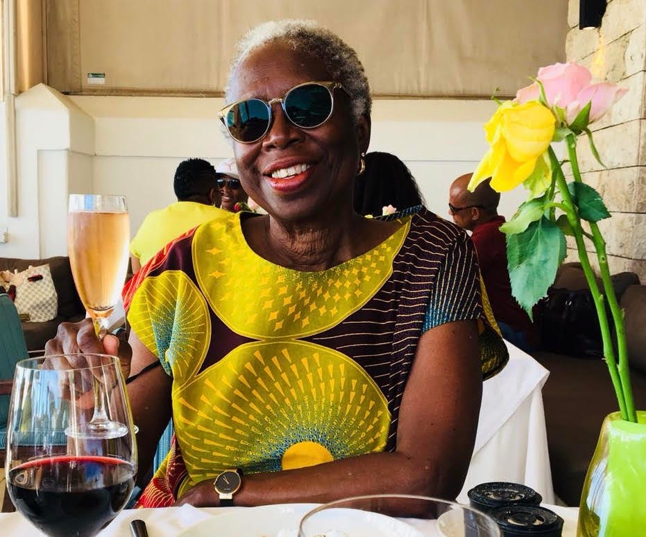 Color photo of the writer Karen Hill Anton wearing sunglasses and an African print dress while seated at a dining room table in a restaurant with a glass raised as if to toast. Color photo of the writer Karen Hill Anton wearing sunglasses and an African print dress while seated at a dining room table in a restaurant with a glass raised as if to toast.