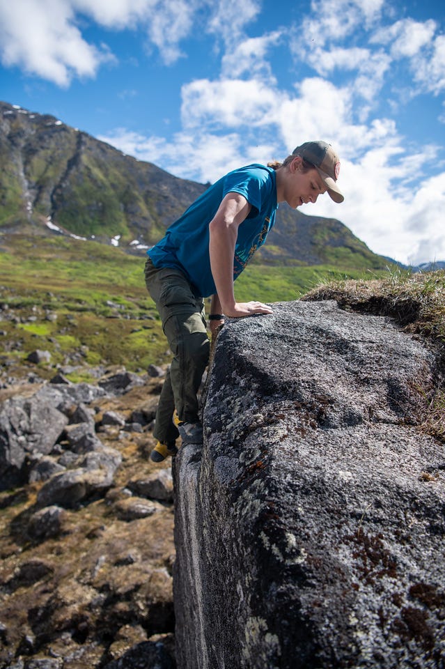 A teenage boy in a blue t-shirt, green pants, and a tan cap presses down with both hands on the rounded top edge of a tall boulder, one foot smeared high on the face as he climbs over the lip, looking down at the rock. Behind him, a green mountainside with a thin waterfall gully rises to the left under a blue sky with bright cumulus. A teenage boy in a blue t-shirt, green pants, and a tan cap presses down with both hands on the rounded top edge of a tall boulder, one foot smeared high on the face as he climbs over the lip, looking down at the rock. Behind him, a green mountainside with a thin waterfall gully rises to the left under a blue sky with bright cumulus.