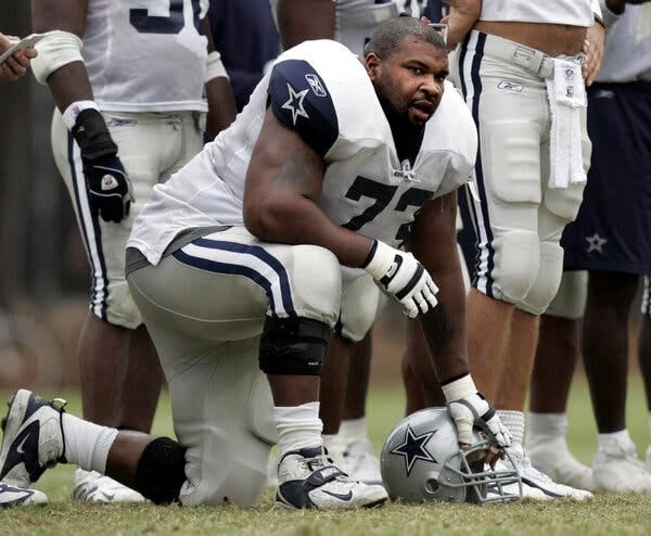 Allen, in uniform, kneels on his left knee, surrounded by teammates. His left hand is on his helmet, which is on the ground in front of him.
