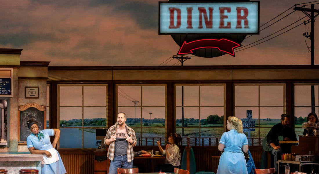 A stage with a diner set, with two waitresses looking at a man pulling his shirt down A stage with a diner set, with two waitresses looking at a man pulling his shirt down