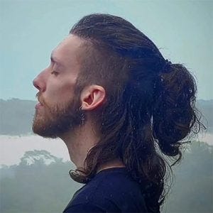 Um homem de perfil com cabelo comprido e preso e expressão neutra. Ele está de olhos fechados, usa barba curta e camiseta escura.