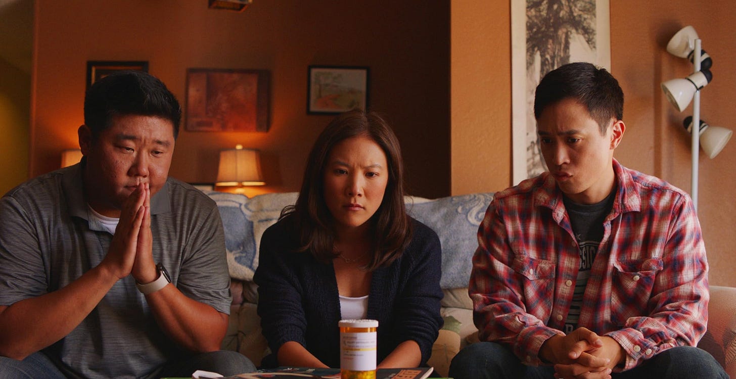 Screenshot of three Asian American siblings sitting on a couch looking at a prescrption pill bottle.