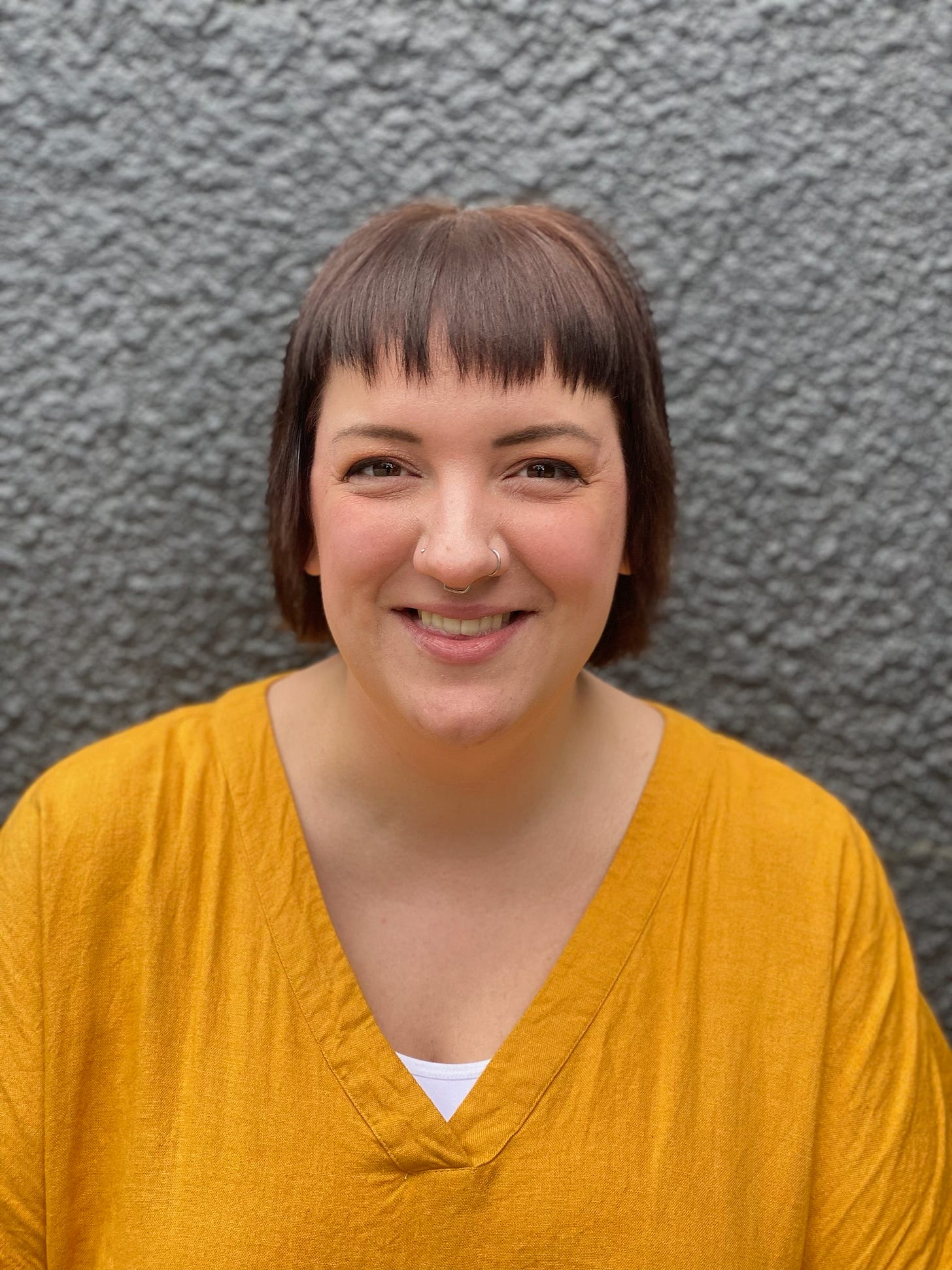 Headshot of Meg Ellard, a woman with a short brown bob and fringe, wearing a mustard yellow top, smiling warmly against a grey textured wall.