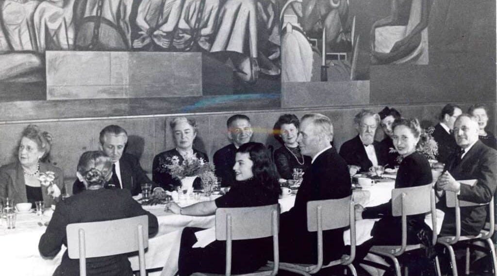 Black and white photo of men and women in 1940s dress seated around a banquet table in hard-back chairs beneath a mural