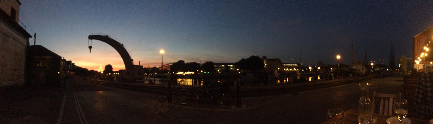Bristol harbourside at sunset from the Olive Shed restaurant with a dockland crane in the background and many lightbulbe glowing above the seating area