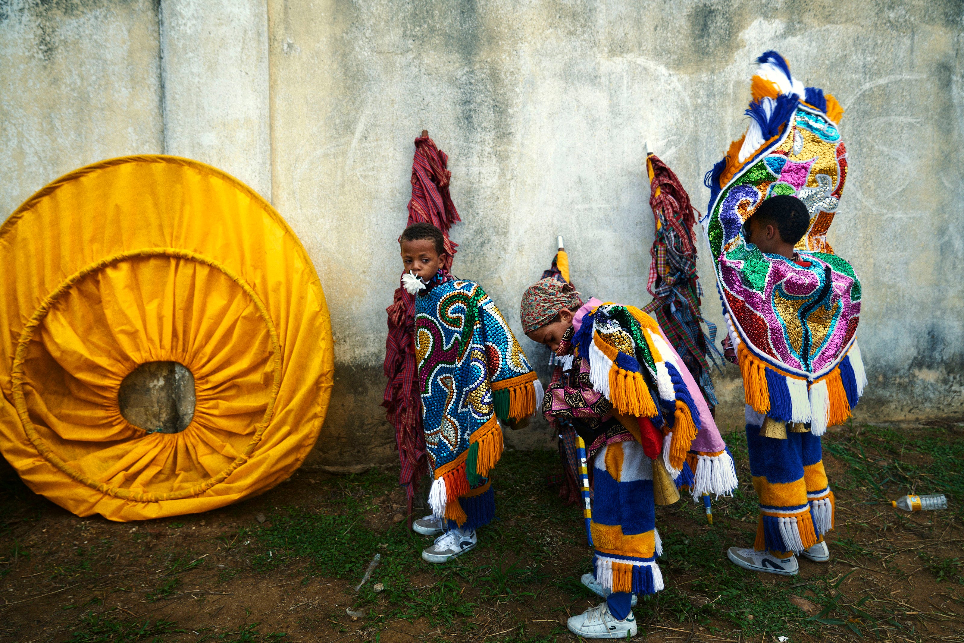 Three children in vibrant, colorful costumes stand near a worn, yellow circular prop and a concrete wall. Two children look down while one gazes forward, all appearing to prepare for a performance. Grass and dirt cover the ground.