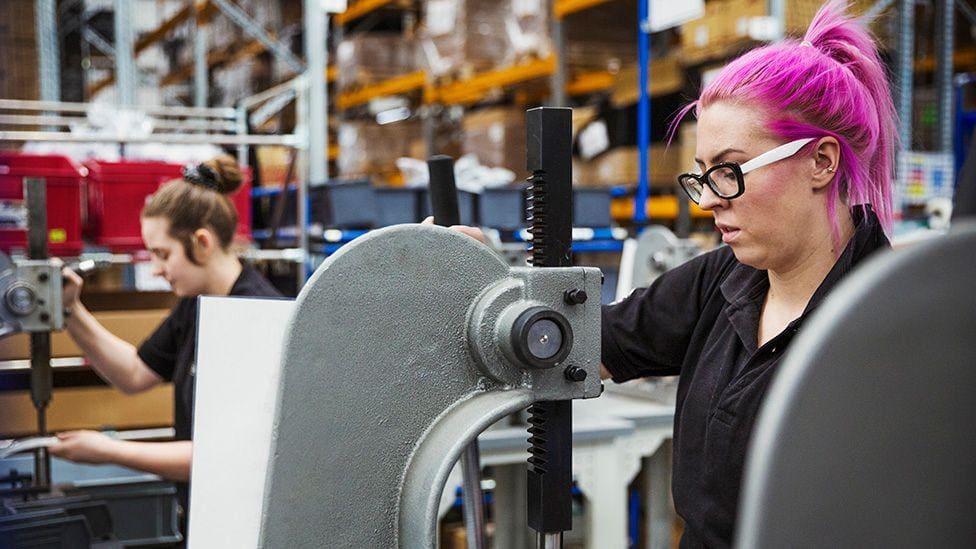 A woman with pink hair uses a hole punch machine in a factory making bicycles, with another worker in the background in a manufacturing environment. A woman with pink hair uses a hole punch machine in a factory making bicycles, with another worker in the background in a manufacturing environment.