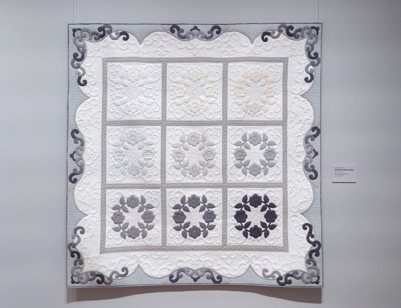 a quilt in shades of white, gray, and black. There is a nine-block pattern with a circular floral pattern in each block. The patterns' shades go from white to black from the top left to the bottom right a quilt in shades of white, gray, and black. There is a nine-block pattern with a circular floral pattern in each block. The patterns' shades go from white to black from the top left to the bottom right
