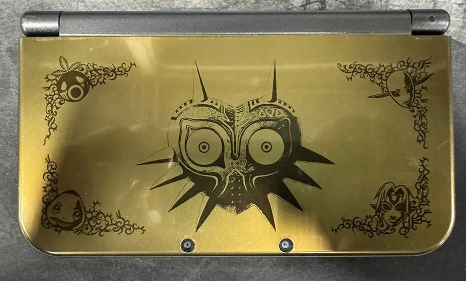 Gold handheld gaming device with intricate black designs featuring a central mask motif and character faces on a textured surface.