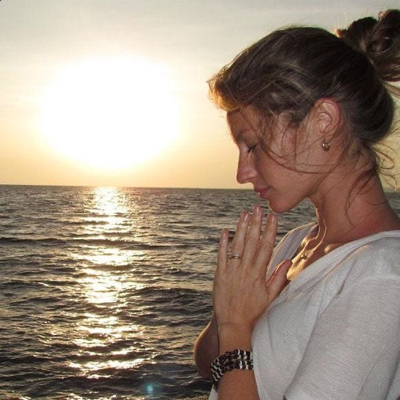 This may contain: a woman standing in front of the ocean with her hands clasped to her chest as the sun sets