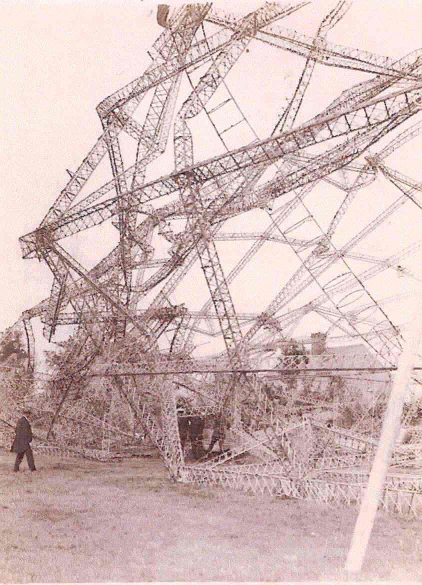 Officers and locals inspect the wreckage at the nose of the zeppelin. Masses of twisted metal towers above them and continues out of shot.