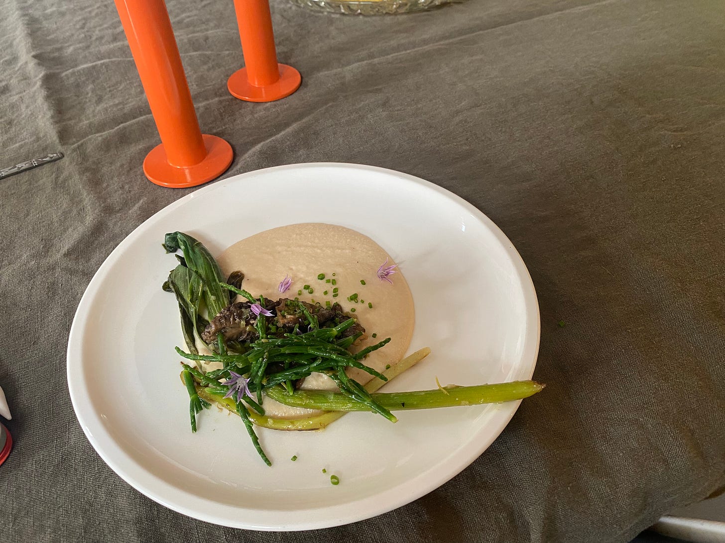 On a white plate, Neil's dish described above: green shoots and mushrooms on a bed of the tofu-onion cream, with chive blossom garnish. The table is laid with a grey tablecloth.