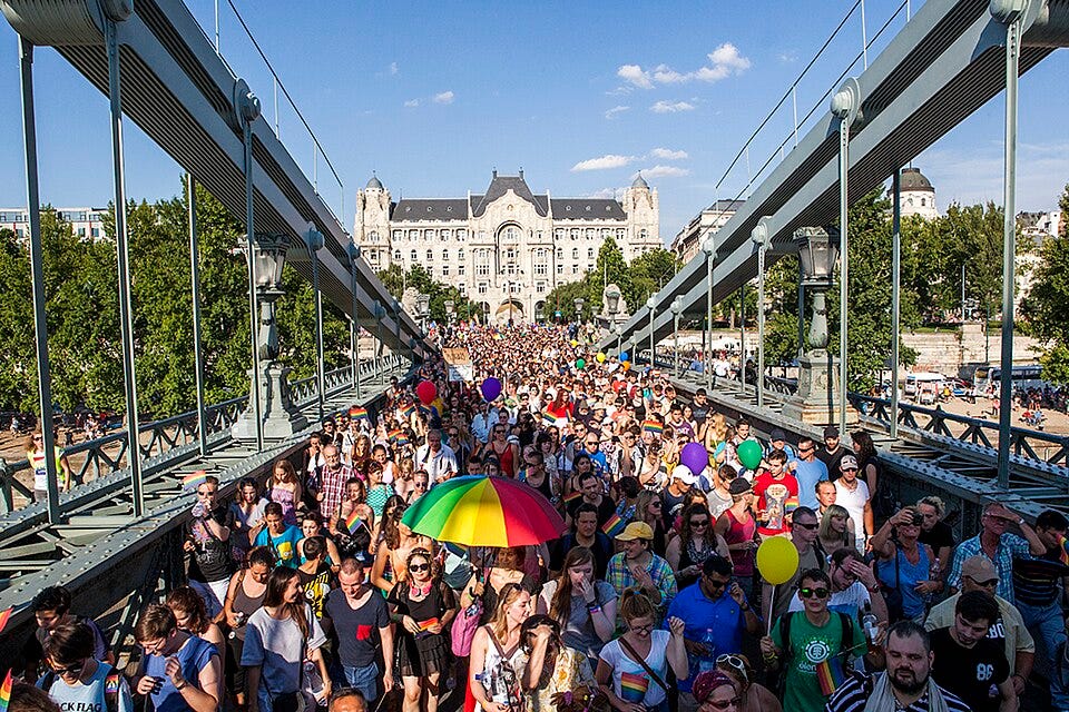 A large crowd marches across Budapest’s Chain Bridge during Budapest Pride, carrying rainbow flags and banners in front of the city skyline.