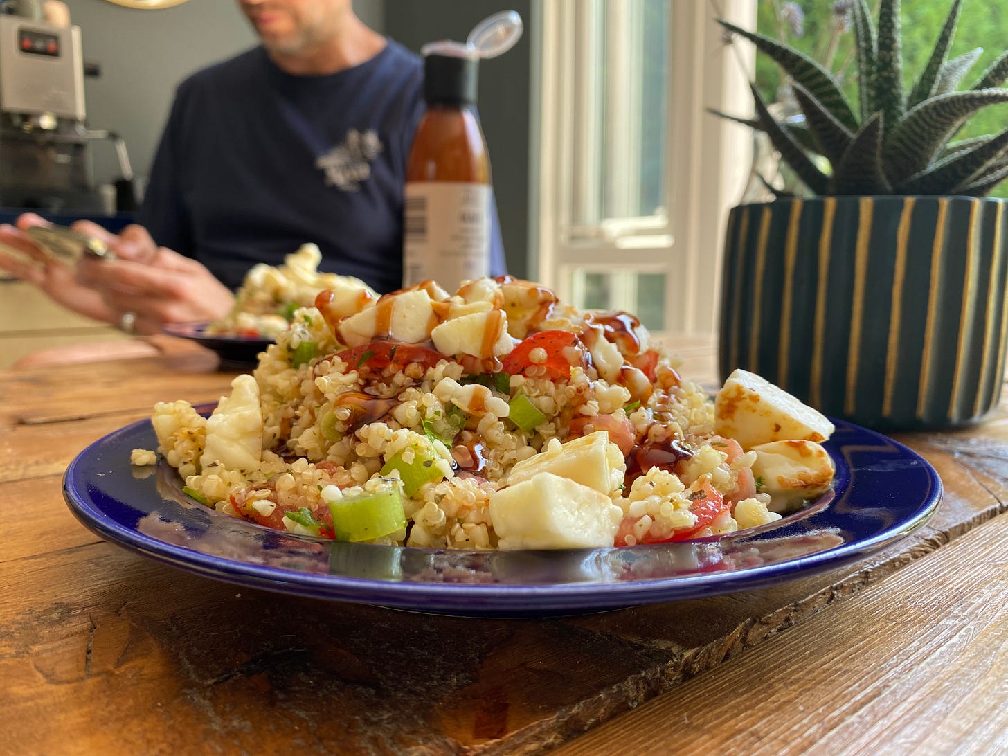 Blue plate holding a mixed grain and vegetable dish, with cubes of cheese on top. A bottle is placed behind the plate. A person is sitting at the table looking at their phone.