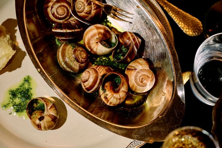 Escargot in garlic and parsley butter served in a silver dish with shells nestled in vibrant green sauce, accompanied by crusty bread and vintage flatware.