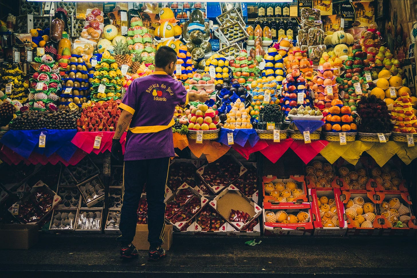 Fotografia em cores de vendedor, de costas, diante de uma barraca de feira cheia de frutas. Fotografia em cores de vendedor, de costas, diante de uma barraca de feira cheia de frutas.