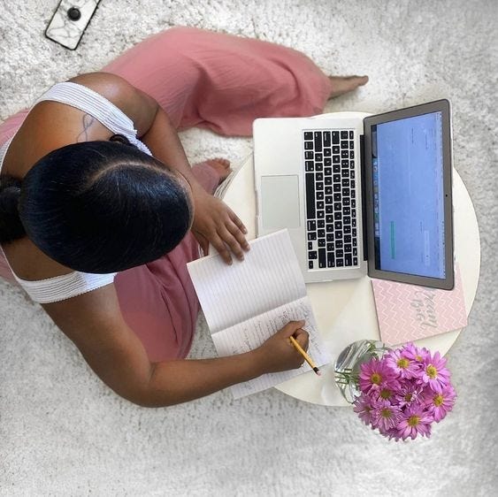 This may contain: a woman sitting at a table writing in front of a laptop computer on top of a white rug