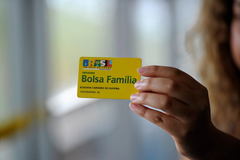 A hand holds a yellow Bolsa Família card in focus, with the cardholder blurred in the background.