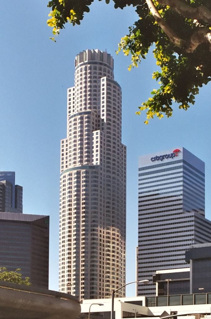 A tall office building with a distinctive shape in an urban setting, surrounded by trees and other buildings.