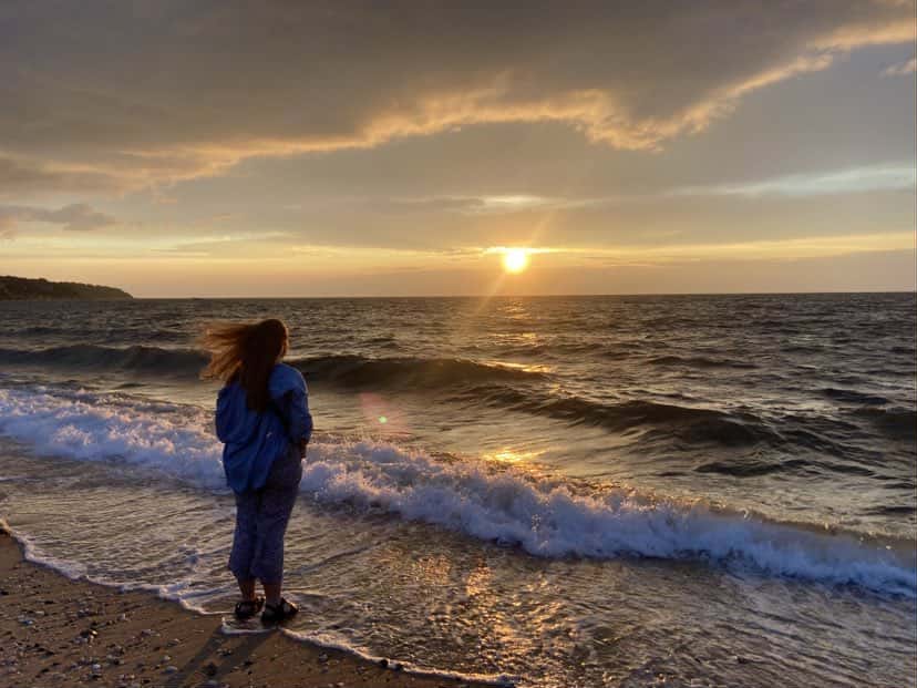 A person stands at the edge of the beach during sunset, facing the ocean waves. The sky is partly cloudy with golden sunlight reflecting across the water, creating a peaceful and contemplative scene.