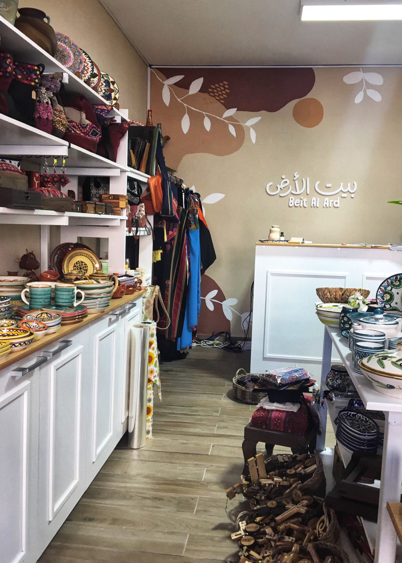 Interior of Beit al-Ard shop in al-Bireh, West Bank. White shelving displays colorful Palestinian ceramics, embroidered bags, wooden crafts, and handmade goods. Traditional embroidered garments hang along the back wall. The shop name appears in raised white lettering in Arabic and English — "Beit Al Ard" — against a warm illustrated mural of leaves and organic shapes.