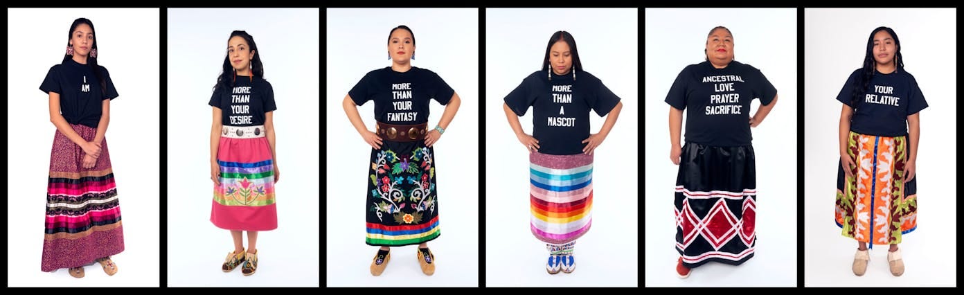 Six indigenous women stand in front of white backgrounds in a horizontal row. each wears a colorful skirt, some wear moccasins, each also wears a black t shirt with white text. In order from left to right they read: I AM, MORE THAN YOUR DESIRE, MORE THAN YOUR FANTASY, MORE THAN A MASCOT, ANCESTRAL LOVE PRAYER SACRIFICE, YOUR RELATIVE Six indigenous women stand in front of white backgrounds in a horizontal row. each wears a colorful skirt, some wear moccasins, each also wears a black t shirt with white text. In order from left to right they read: I AM, MORE THAN YOUR DESIRE, MORE THAN YOUR FANTASY, MORE THAN A MASCOT, ANCESTRAL LOVE PRAYER SACRIFICE, YOUR RELATIVE