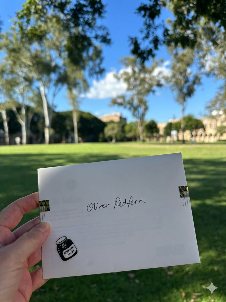 Envelope held up in afternoon sunlight outdoors, addressed "Oliver Redfern" in handwriting, with a small illustrated ink bottle stamp. A green lawn and eucalyptus trees are visible out of focus in the background.