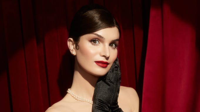 Dylan Mulvaney poses in front of a red velvet curtain.