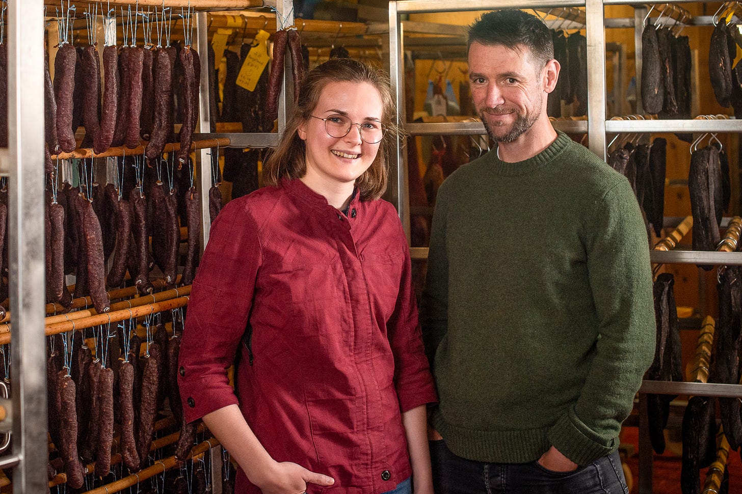 Davina Sialm und Adrian Hirt stehen im Reiferaum für Trockenfleisch. Links Davina Sialm in rotem Arbeitshemd und mit Brille, rechts Adrian Hirt im grünen Pullover. Beide lächeln in die Kamera. Hinter ihnen hängen dicht an dicht dunkle Würste und Fleischstücke an Holzstangen und Metallgestellen.