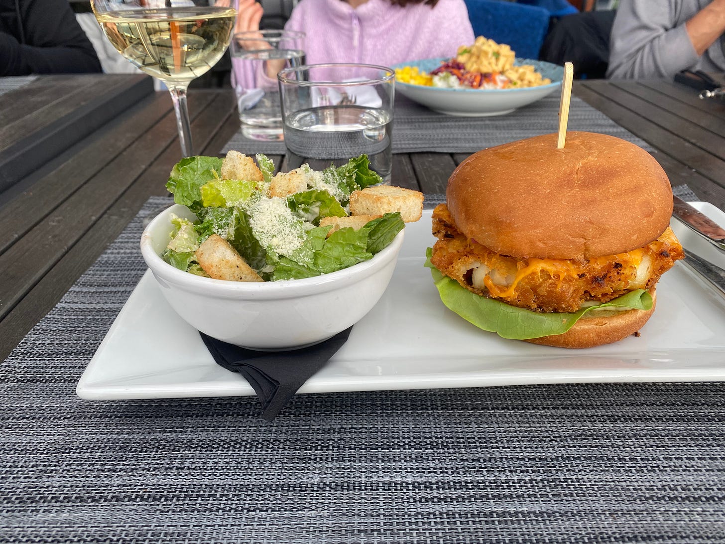 A rectangular white plate with a small bowl of caesar salad on the left, a fried fish sandwich on the right. The sandwich is on a brioche bun with lettuce and cheese. Behind the plate is a glass of white wine, and a young girl in a purple sweater is just visible across the table sitting in front of a blue bowl of vegetables and tofu.