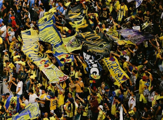 A large crowd of soccer fans fills a stadium section, waving yellow and blue flags and banners with the Mexican Club América emblem in celebration.