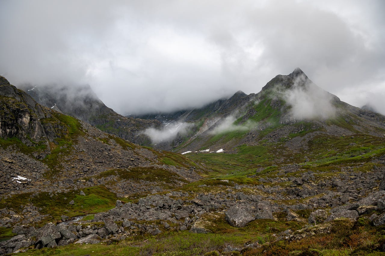 A view up a green tundra valley strewn with grey boulders, low brush in the foreground still dark with rain. A sharp rocky peak rises at right, its lower slopes green, while heavy white cloud pours over the ridgeline and drifts in patches partway down the mountainsides, hiding the valley's head.