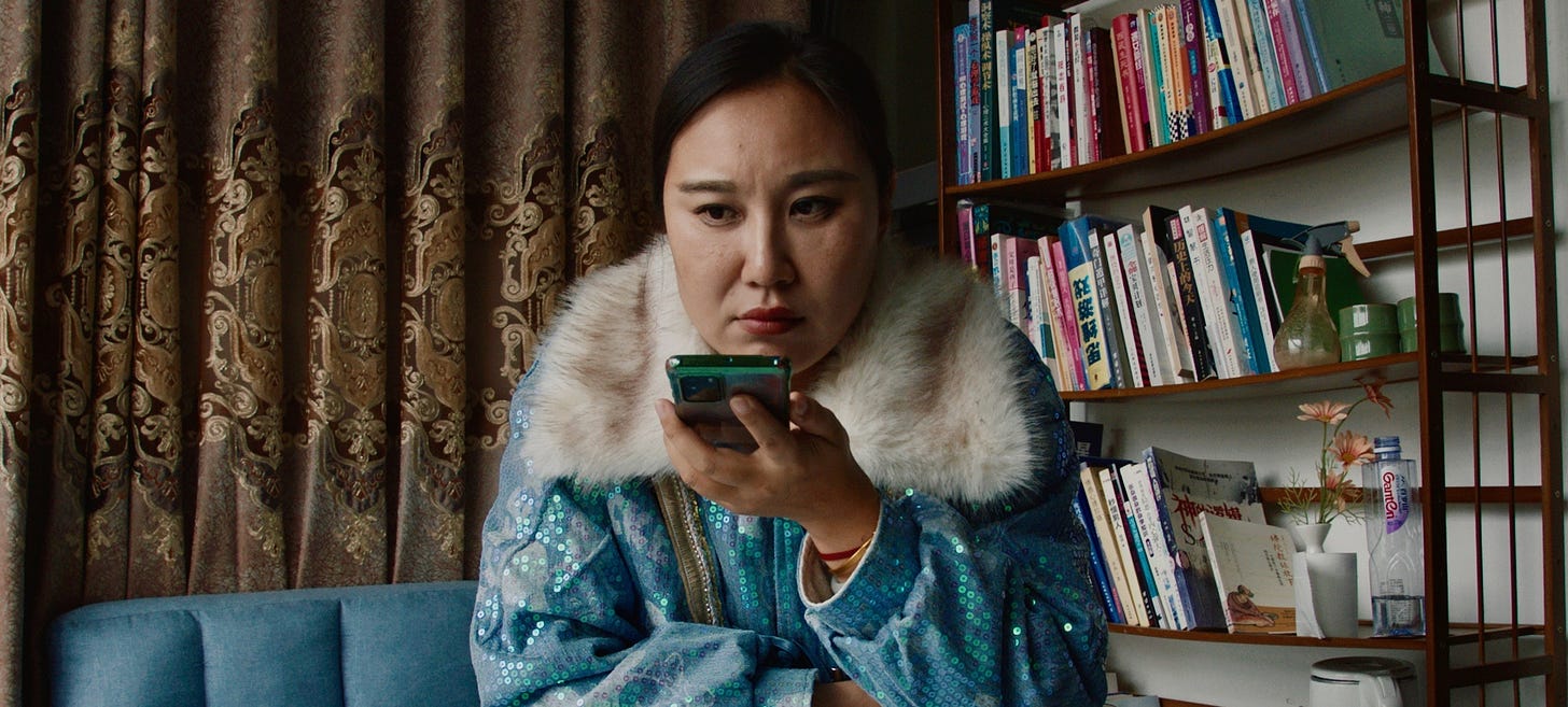 A person wearing a sparkly jacket with a fur collar speaks into a phone. They are seated at a table in front of a bookshelf.