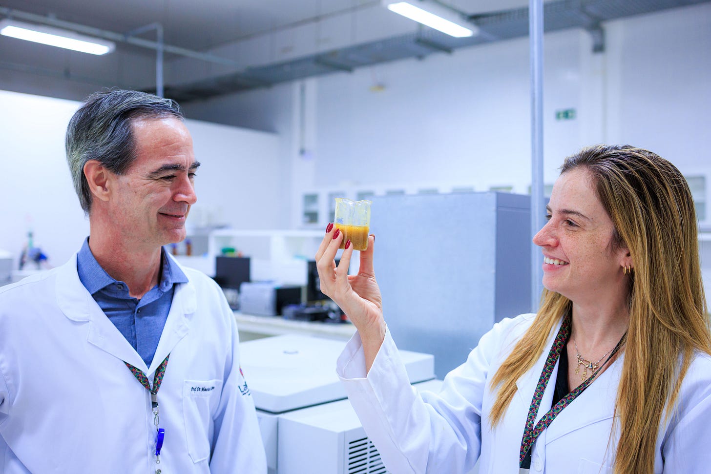 Fotografia em ambiente de laboratório exibe dois adultos vestindo jalecos brancos e cordões coloridos, sorridentes, observando um béquer contendo substância de coloração amarelo-alaranjada erguido pela mulher à direita, de cabelos castanhos compridos e unhas vermelhas. O homem à esquerda, de cabelos grisalhos, veste jaleco com identificação parcialmente visível. Ao fundo, desfocados, são visíveis equipamentos e bancadas de laboratório. Fim da descrição. Fotografia em ambiente de laboratório exibe dois adultos vestindo jalecos brancos e cordões coloridos, sorridentes, observando um béquer contendo substância de coloração amarelo-alaranjada erguido pela mulher à direita, de cabelos castanhos compridos e unhas vermelhas. O homem à esquerda, de cabelos grisalhos, veste jaleco com identificação parcialmente visível. Ao fundo, desfocados, são visíveis equipamentos e bancadas de laboratório. Fim da descrição.