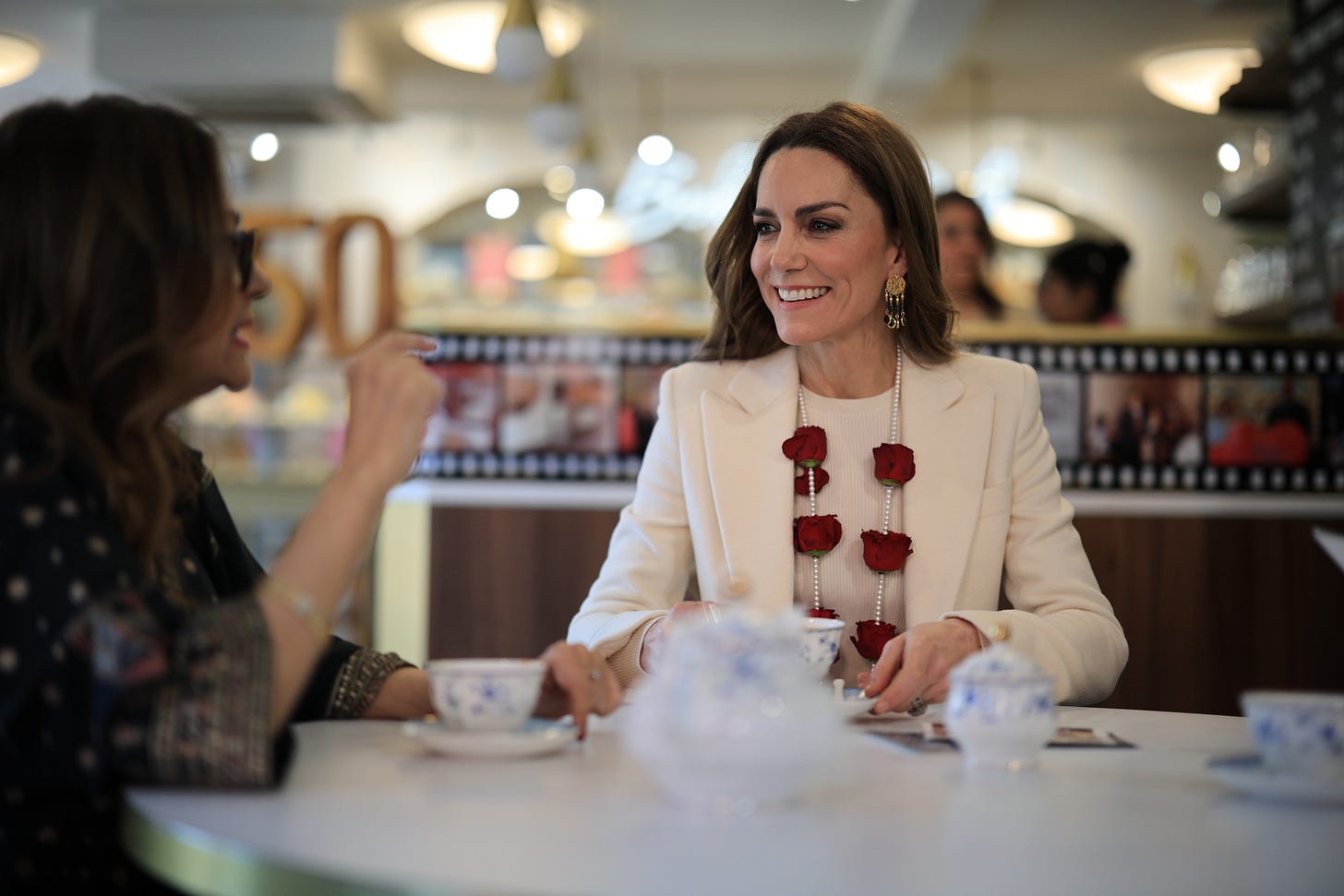 Princess Kate wearing a white suit with a garland of flowers sitting at a table smiling having a cup of tea