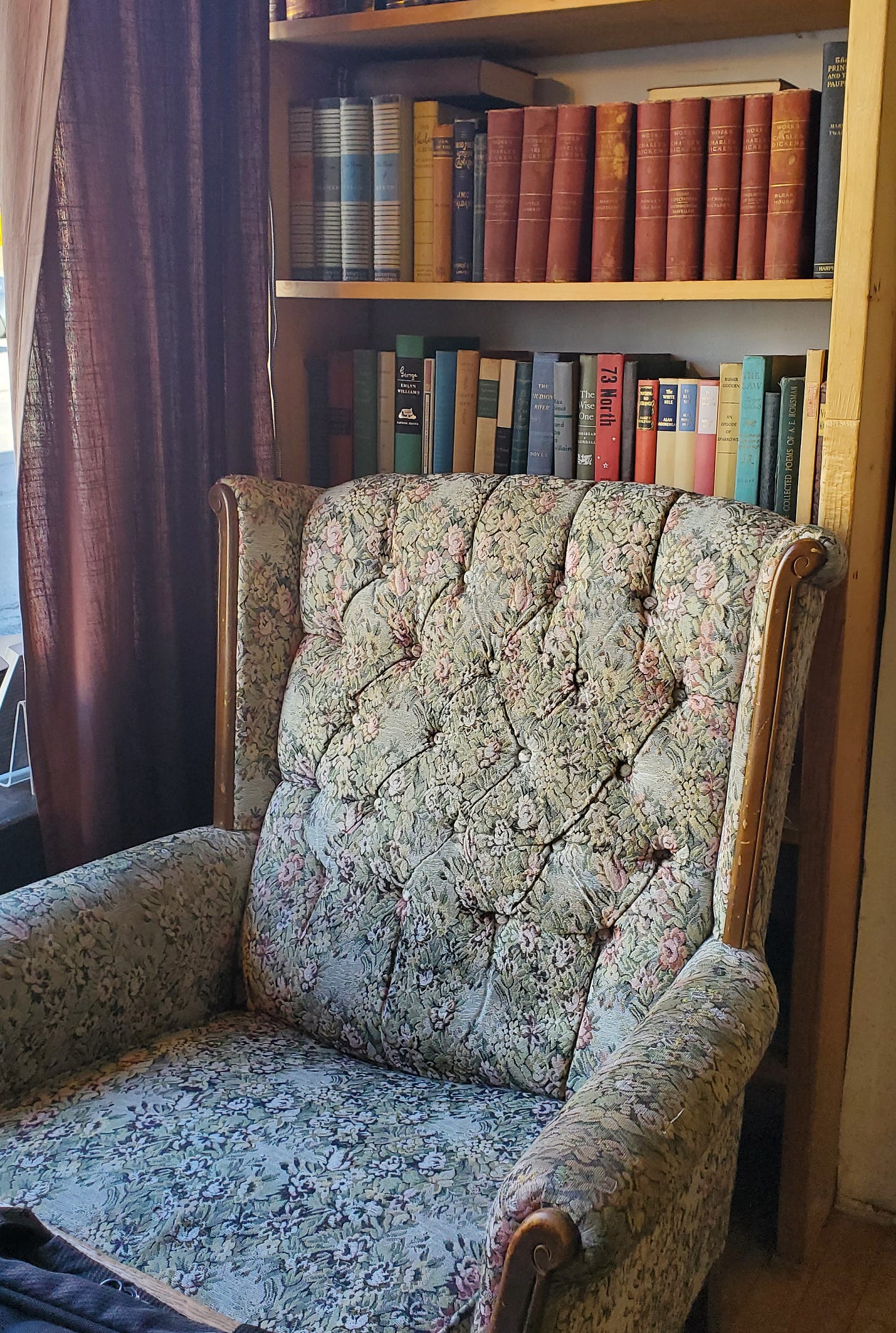 A comfy chair with wooden embellishments and a small floral pattern sits in front of a bookshelf filled with old looking books.