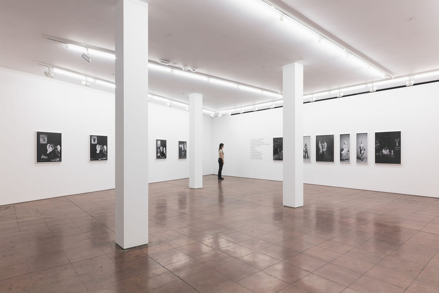 A person stands in a modern, spacious exhibition gallery with polished brown floors and bright lighting, viewing black-and-white photographs on crisp white walls. There are 3 white columns throughout the room, and there are 10 visible works on the wall. On the far right wall, there is a block of black text next to the start of some evenly spaced works on the wall.