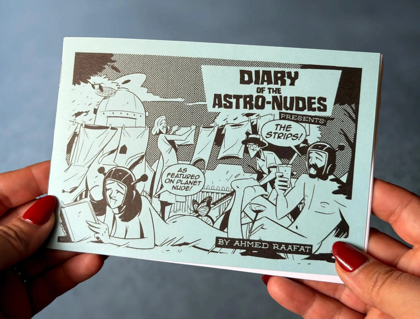A hand holds the blue cover of Diary of the Astro-Nudes Presents: The Strips! by Ahmed Raafat. The illustration shows Arr and Arreya lounging nude in a backyard scene, surrounded by laundry on a clothesline, a saucer-like ship, and a group of surprised onlookers. A speech bubble on the cover reads, “As featured on Planet Nude!”