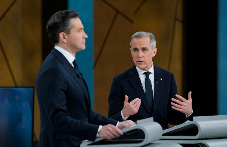 One man with short grey hair on a stage gestures as he speaks while looking over at a dark-haired man staring straight ahead. One man with short grey hair on a stage gestures as he speaks while looking over at a dark-haired man staring straight ahead.